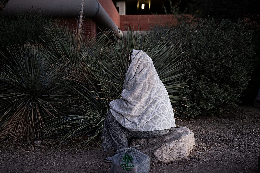 An unhoused person wrapped in blanket and sitting on a rock outdoors in Tucson, Arizona.